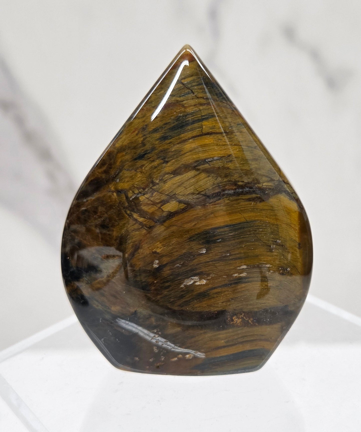 A polished tiger's eye gemstone cabochon displayed on a clear acrylic stand against a white marble background.