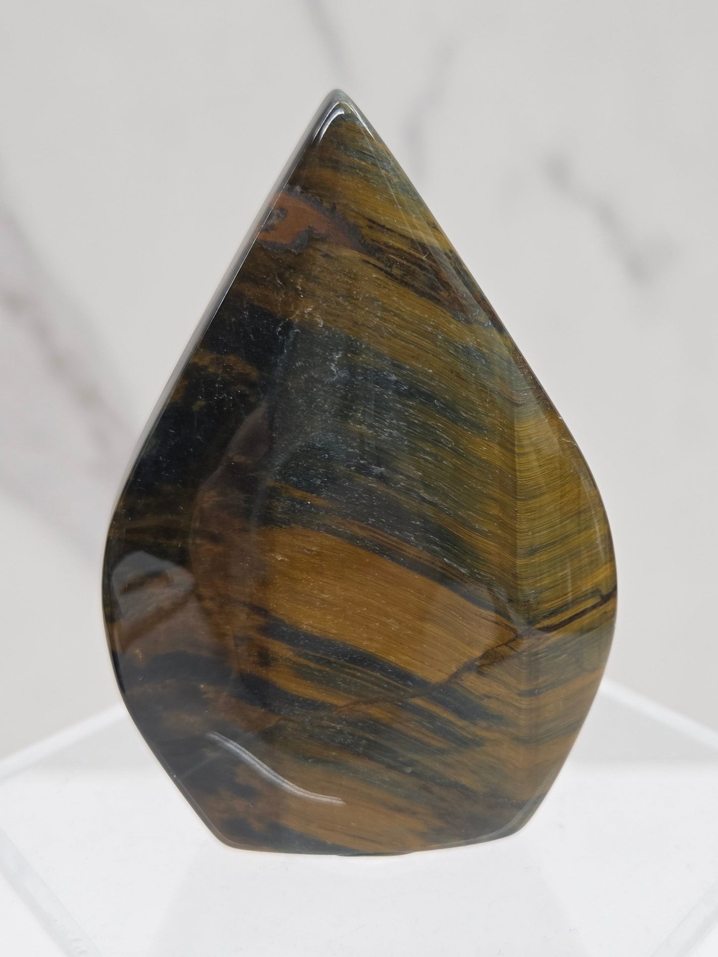 A polished tiger's eye stone cabochon with characteristic golden-brown and black banded striations, displayed on a clear acrylic stand against a white marble background.