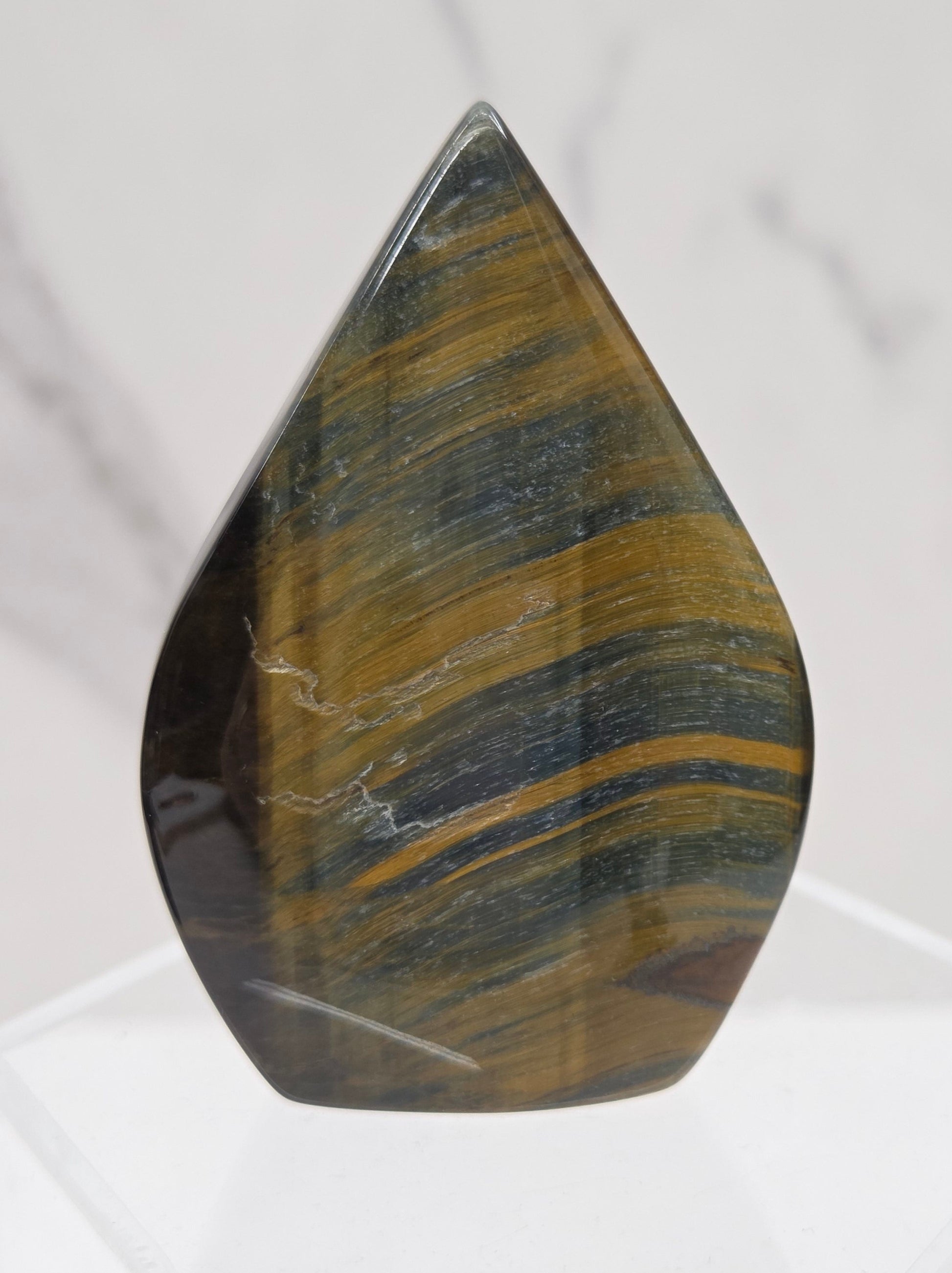 A polished tiger's eye stone chip with characteristic golden-brown and dark green banded striations, displayed on a clear acrylic stand against a light marble background.