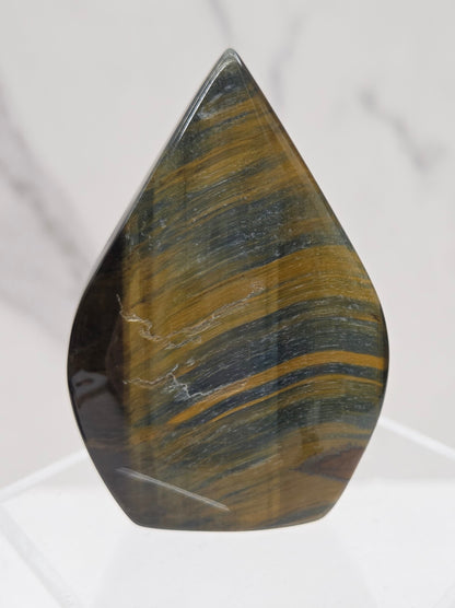 A polished tiger's eye stone chip with characteristic golden-brown and dark green banded striations, displayed on a clear acrylic stand against a light marble background.