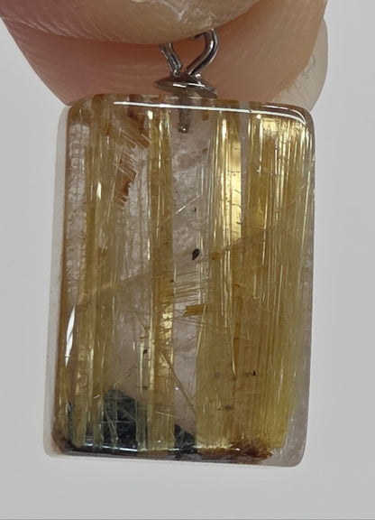 A rectangular clear gemstone pendant with prominent golden-yellow needle-like inclusions, held by fingers against a light gray background.