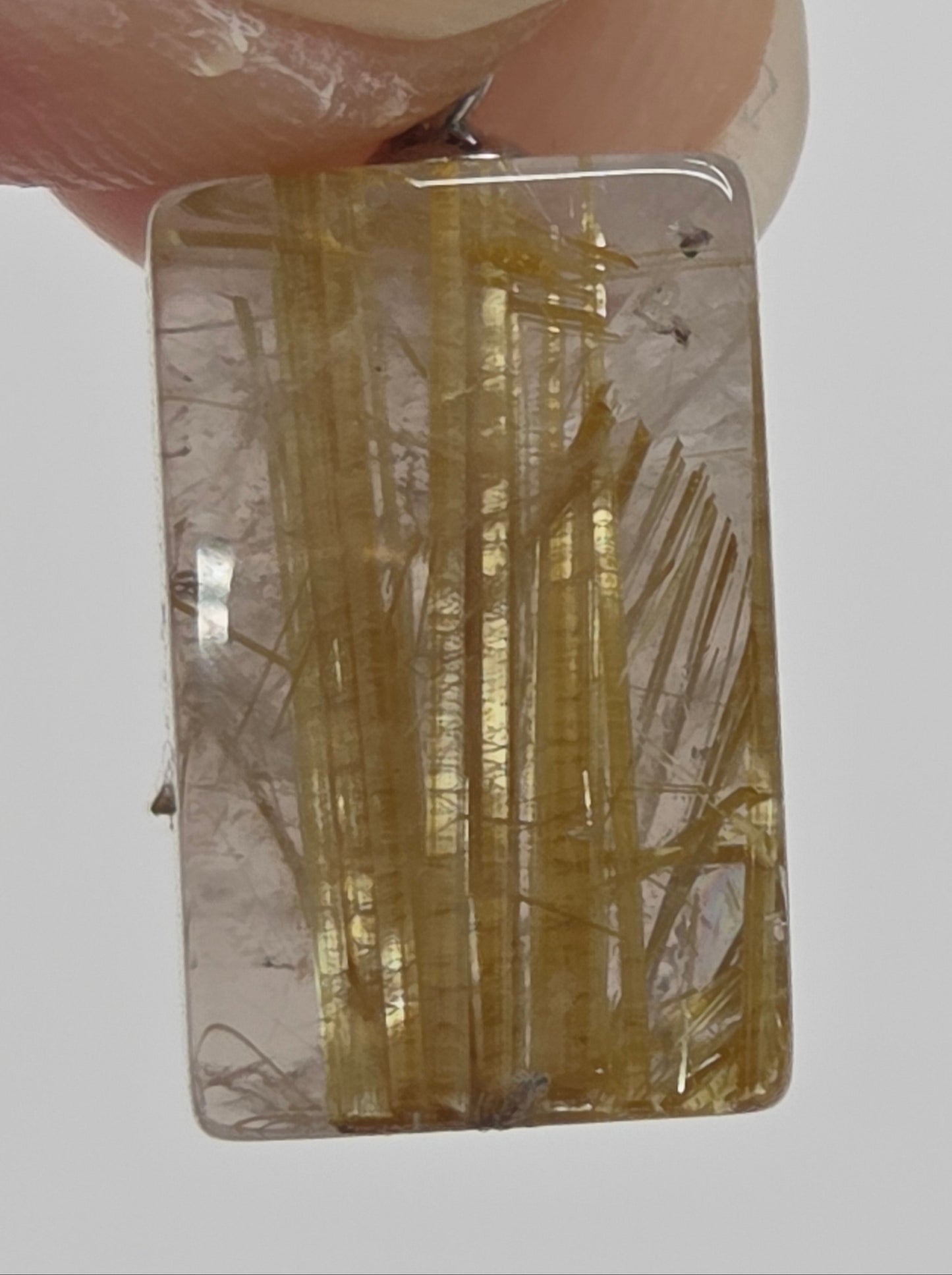 A close-up of a rectangular clear resin or glass pendant with embedded golden-yellow hair-like inclusions, held between fingers against a light gray background.
