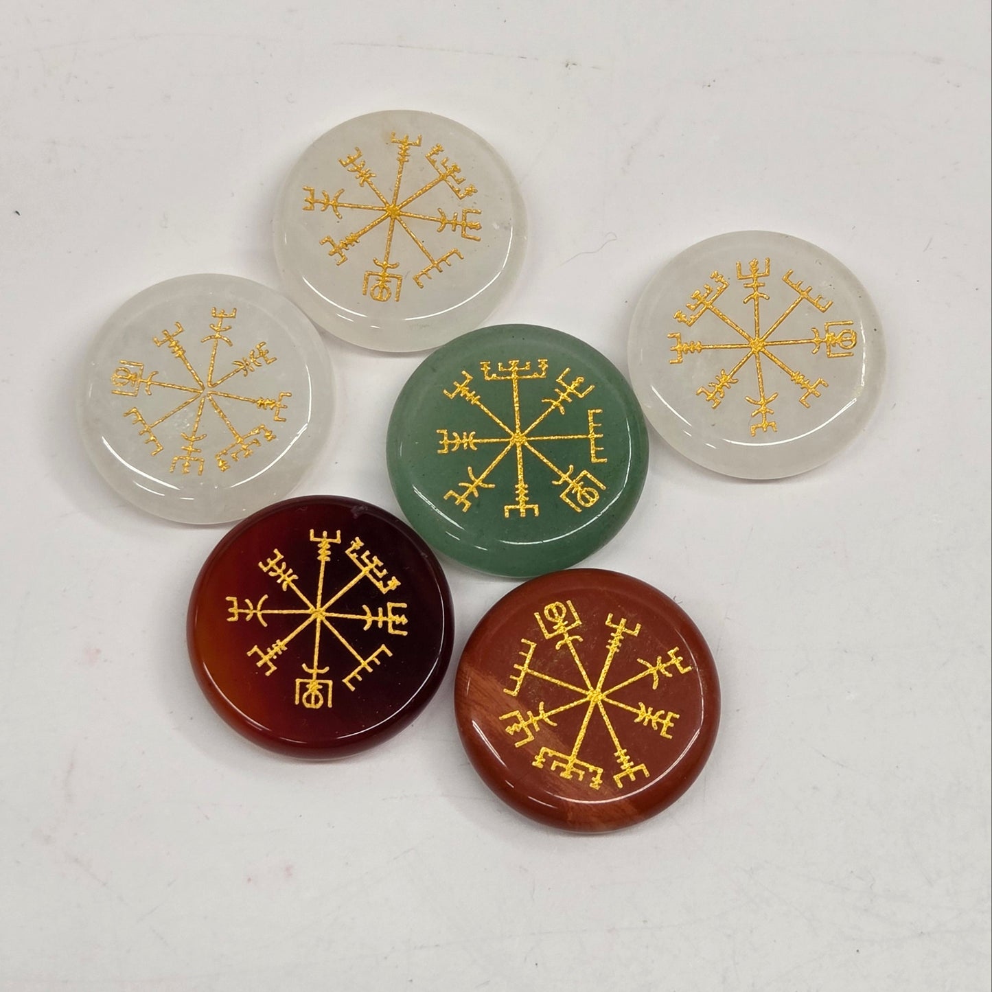 Six round cabochon stones with gold compass rose engravings, arranged on a light off-white background. The stones are translucent white, green, and translucent reddish-brown.