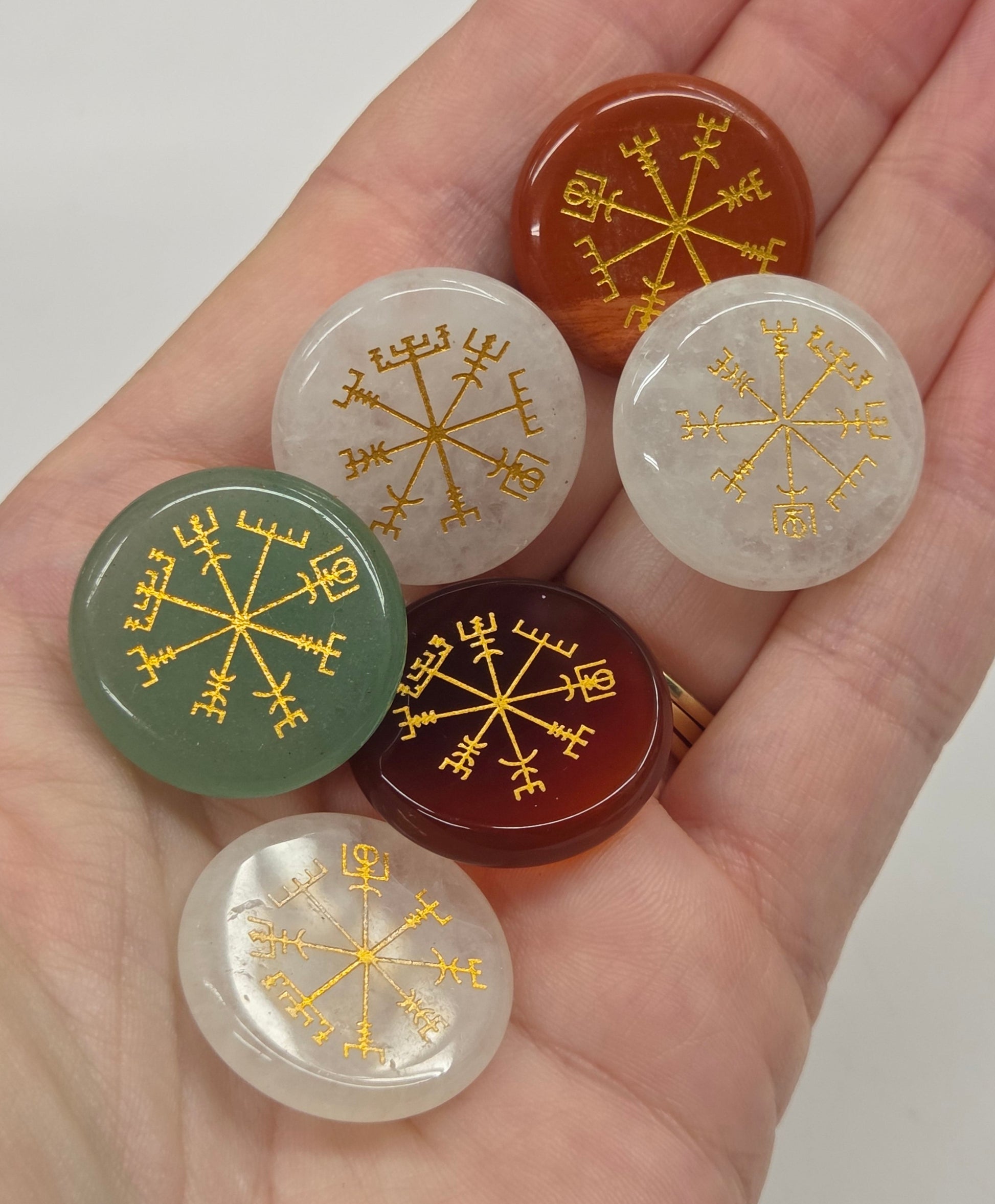 Six round cabochon gemstone pendants with gold Norse runes and compass motifs, held in a person's hand to show scale.