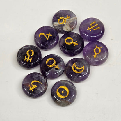 Nine polished purple amethyst gemstone cabochons, each engraved with a different astrological symbol, arranged on a plain white surface.