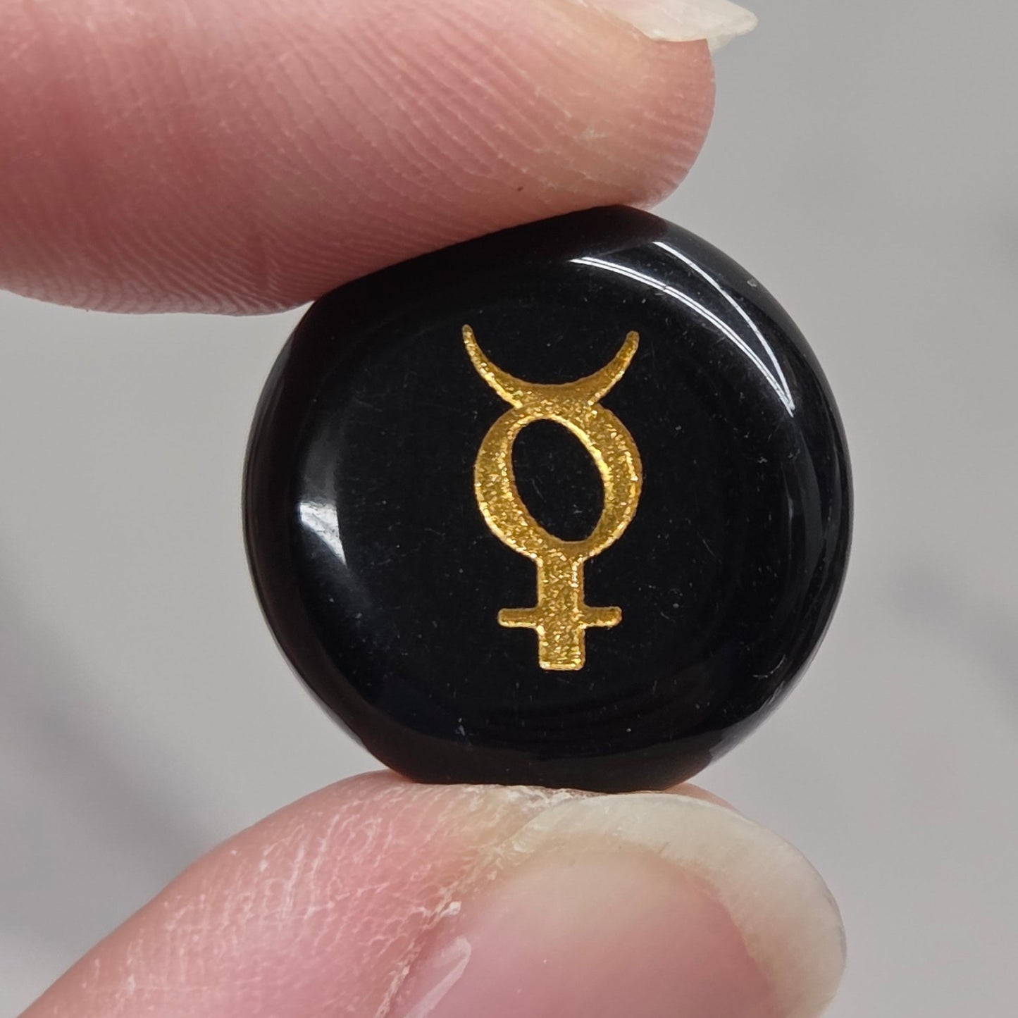 A close-up of a person's fingers holding a small, round, glossy black stone or cabochon featuring a gold astrological symbol, set against a soft, out-of-focus light gray background.