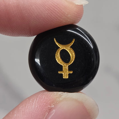 A close-up of a person's fingers holding a small, round, glossy black stone or cabochon featuring a gold astrological symbol, set against a soft, out-of-focus light gray background.