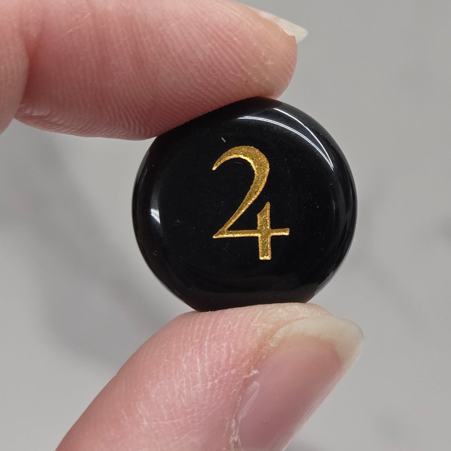 A close-up of a person's fingers holding a small, round, glossy black button or cabochon featuring a gold-colored monogram '24' against a light gray marble background.