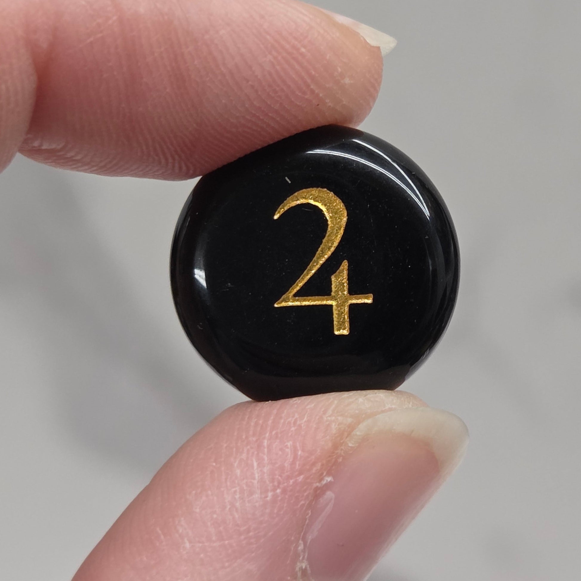A close-up of a person's fingers holding a small, round, glossy black button or cabochon featuring a gold-colored monogram '24' against a light gray marble background.