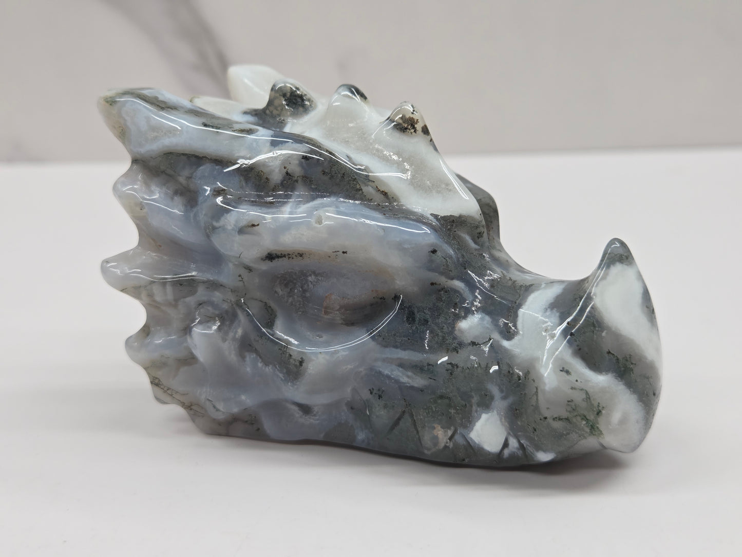 A polished carved stone sculpture resembling a dragon head, featuring natural marbled gray, white, and black patterns, displayed on a neutral light gray surface.
