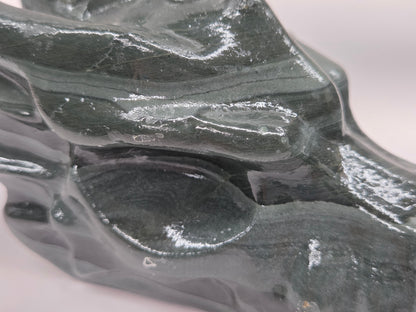 A close-up of a glossy dark gray stone sculpture with organic, flowing contours and reflective highlights on a plain light background.