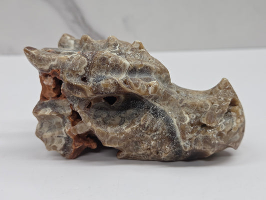 A natural polished stone skull specimen with textured, organic surfaces in earthy gray, beige, and brown tones, displayed on a neutral light gray surface.