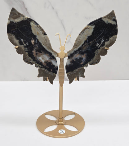 A wall-mounted butterfly-shaped coat rack featuring dark marbled wings in black, gray, and white, with a gold-toned metal mounting bracket and hooks, shown against a white wall and light marble surface.
