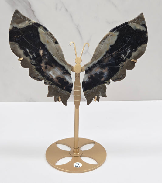 A wall-mounted butterfly-shaped coat rack featuring dark marbled wings in black, gray, and white, with a gold-toned metal mounting bracket and hooks, shown against a white wall and light marble surface.