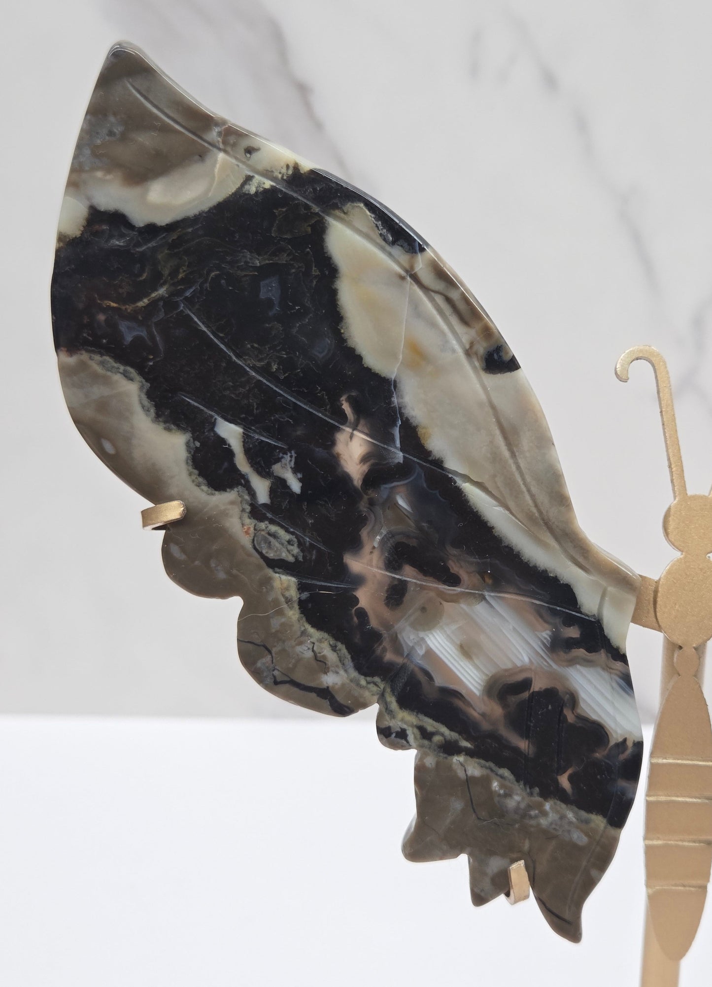 A close-up of a decorative object featuring a large marbled stone wing in black, beige, and gray, attached to a light wood or brass hook-like structure against a white marble background.