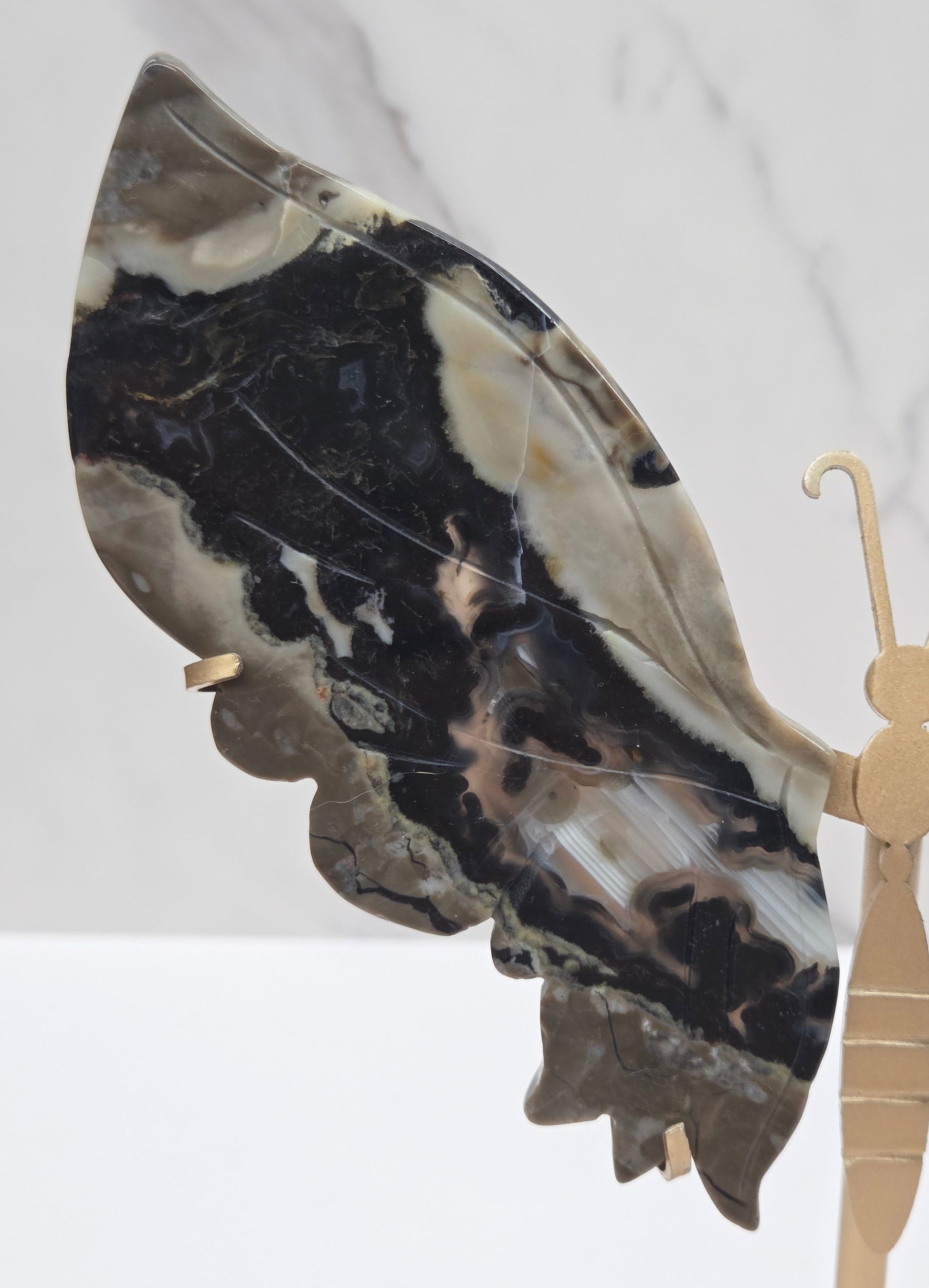 A close-up of a decorative object featuring a large marbled stone wing in black, beige, and gray, attached to a light wood or brass hook-like structure against a white marble background.