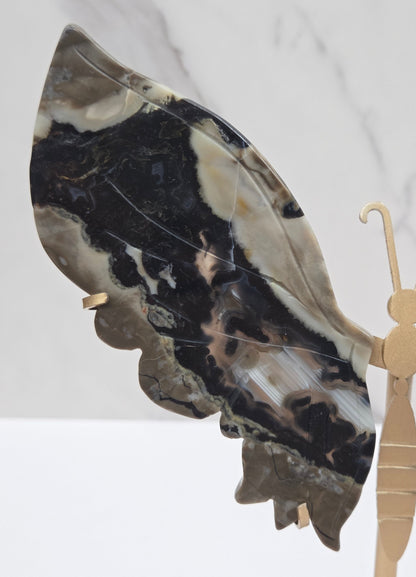 A close-up of a decorative object featuring a large marbled stone wing in black, beige, and gray, attached to a light wood or brass hook-like structure against a white marble background.