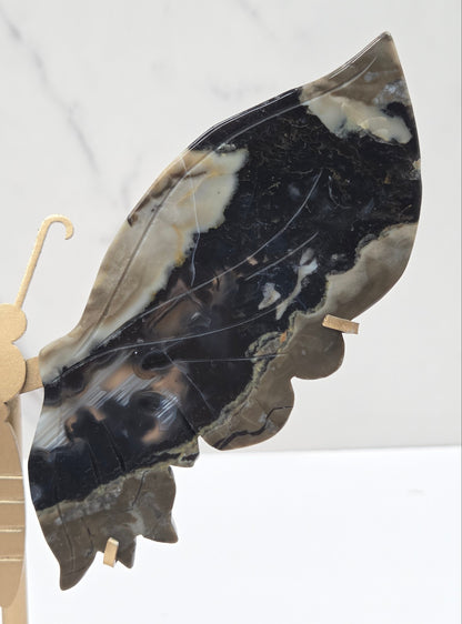 A decorative wall-mounted butterfly sculpture made from polished black, gray, and cream agate stone wings, supported by a matte gold metal frame against a white marble background.