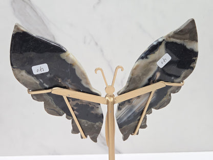 A butterfly-shaped wall decor piece with black and gray marbled wings mounted on a gold metal stand, set against a white marble-patterned background.