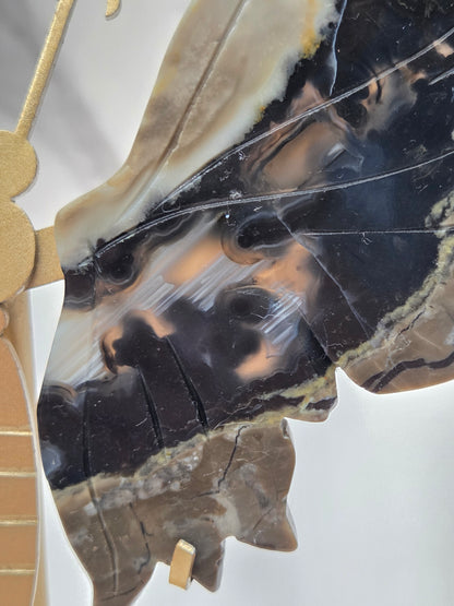 A close-up of a carved stone or agate leaf-shaped object with natural black, brown, cream, and gray banded patterns, photographed against a light neutral background.