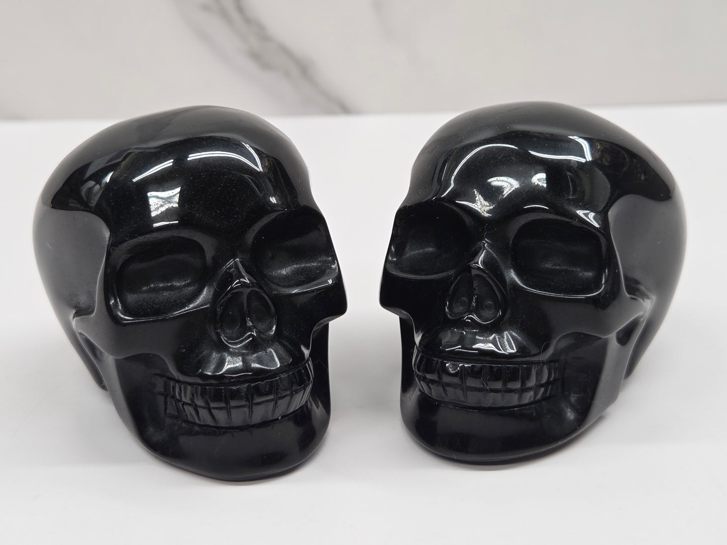 Two glossy black skull-shaped objects displayed side by side on a white surface against a light gray marble-patterned background.