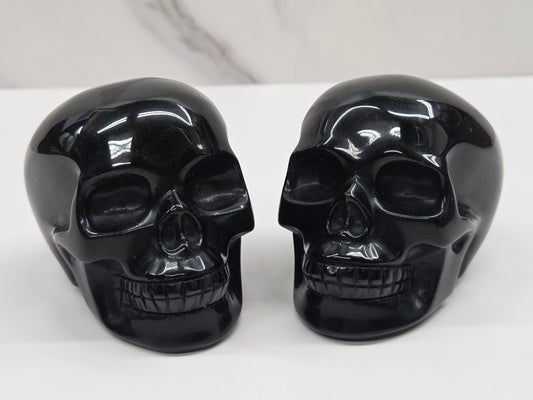 Two glossy black skull-shaped objects displayed side by side on a white surface against a light gray marble-patterned background.