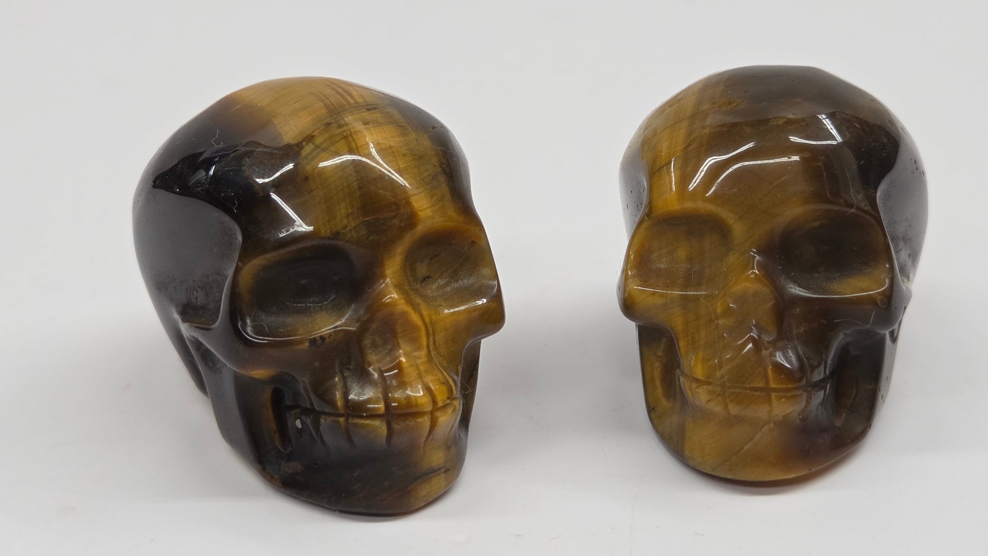 Two polished tiger's eye skull carvings displayed side by side on a plain light gray background.