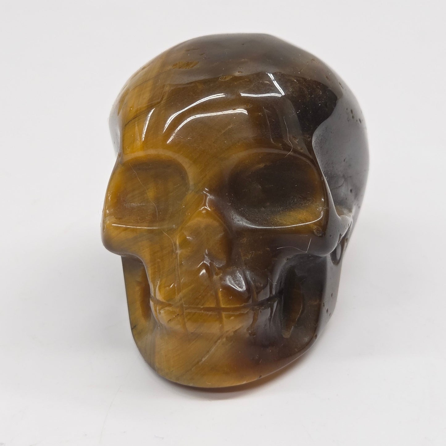 A polished carved skull figurine made of tiger's eye stone, featuring natural brown and black banded patterns, set against a plain white background.