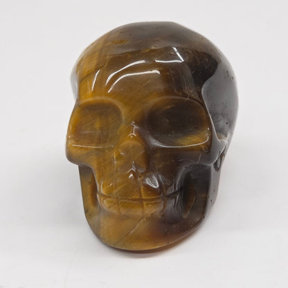 A polished carved skull figurine made of tiger's eye stone, featuring natural brown and black banded patterns, set against a plain white background.