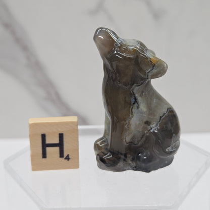 A small carved stone figurine of a seated dog, shown on a clear acrylic stand next to a wooden Scrabble tile with the letter H, against a light gray marble-patterned background.