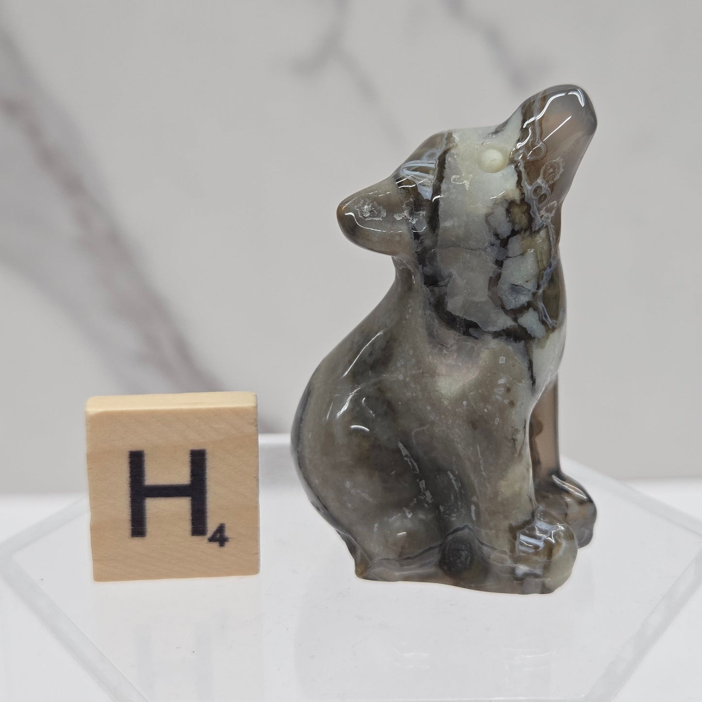 A small carved stone figurine of a seated animal, likely a fox or wolf, shown on a clear acrylic stand next to a wooden Scrabble tile with the letter H4. The figurine has a polished, glossy finish with natural marbled patterns in gray, brown, and off-white.