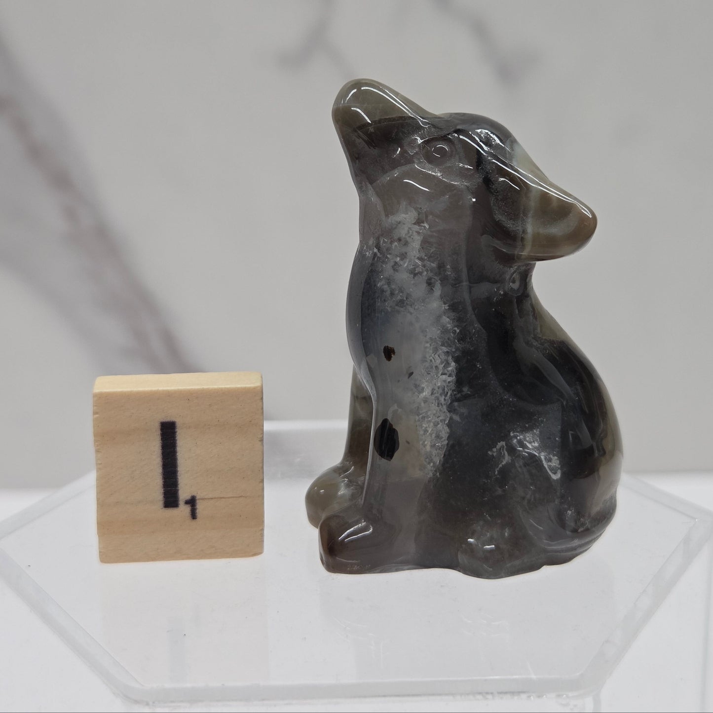 A small carved dog figurine made of dark gray agate or stone, sitting next to a wooden block with the number one on it, displayed on a clear acrylic stand against a light gray marble-patterned background.