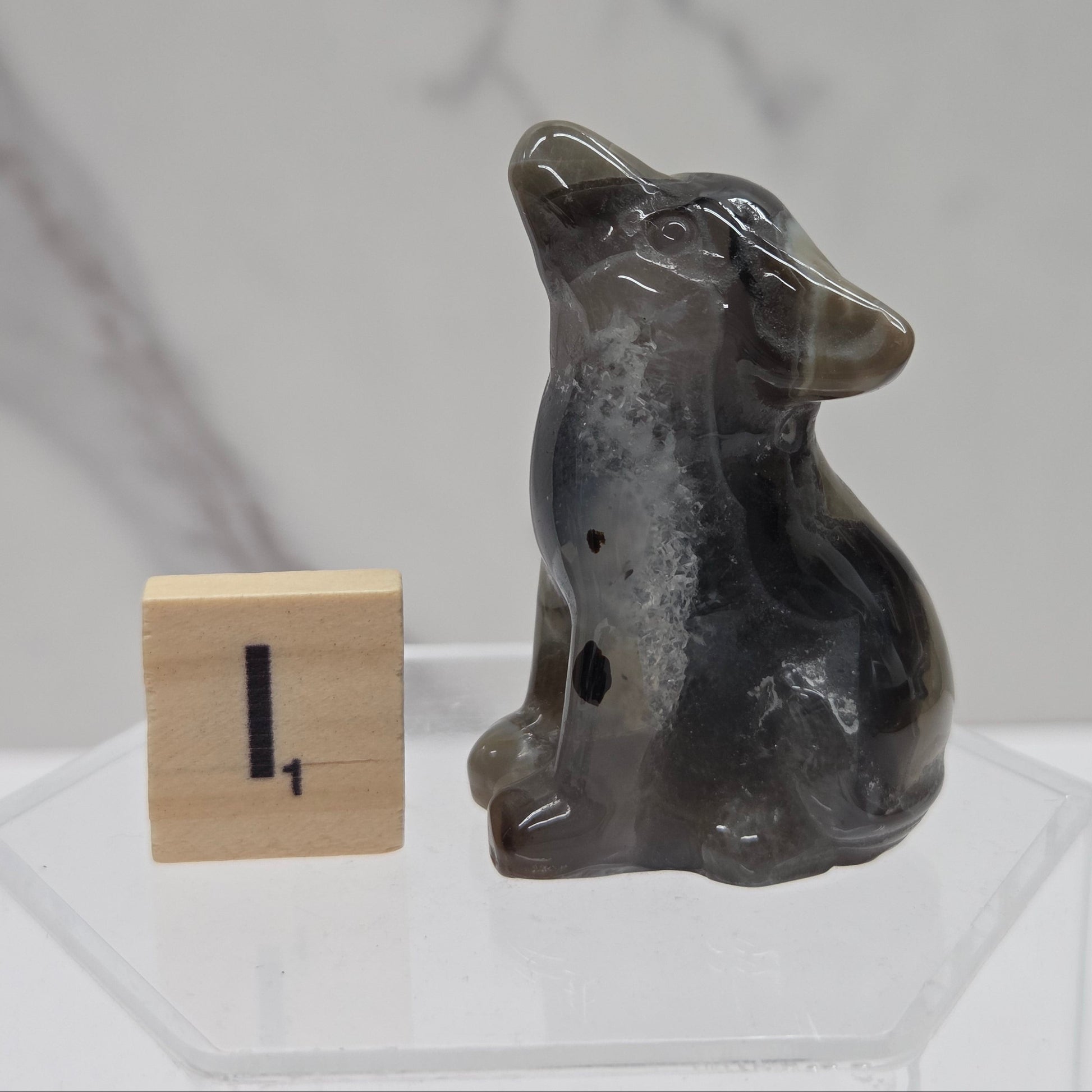 A small carved dog figurine made of dark gray agate or stone, sitting next to a wooden block with the number one on it, displayed on a clear acrylic stand against a light gray marble-patterned background.