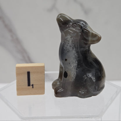 A small carved dog figurine made of dark gray agate or stone, sitting next to a wooden block with the number one on it, displayed on a clear acrylic stand against a light gray marble-patterned background.