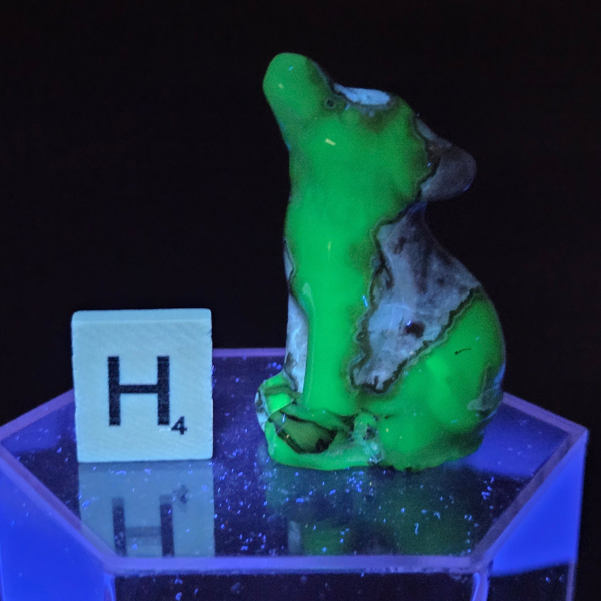 A small sculpted figurine in glowing neon green and gray sits on a translucent blue-lit hexagonal base next to a white tile marked H4 against a black background.