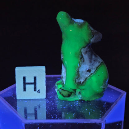 A small sculpted figurine in glowing neon green and gray sits on a translucent blue-lit hexagonal base next to a white tile marked H4 against a black background.