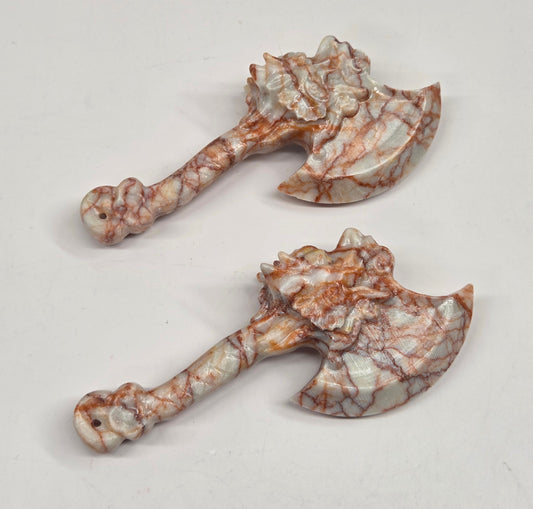 Two small carved axe-shaped ornaments with a white and brown marbled stone pattern, displayed on a plain light background.