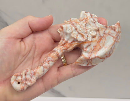 A hand holding a carved decorative axe head made of white stone with orange-brown veining, against a light marble-like background.