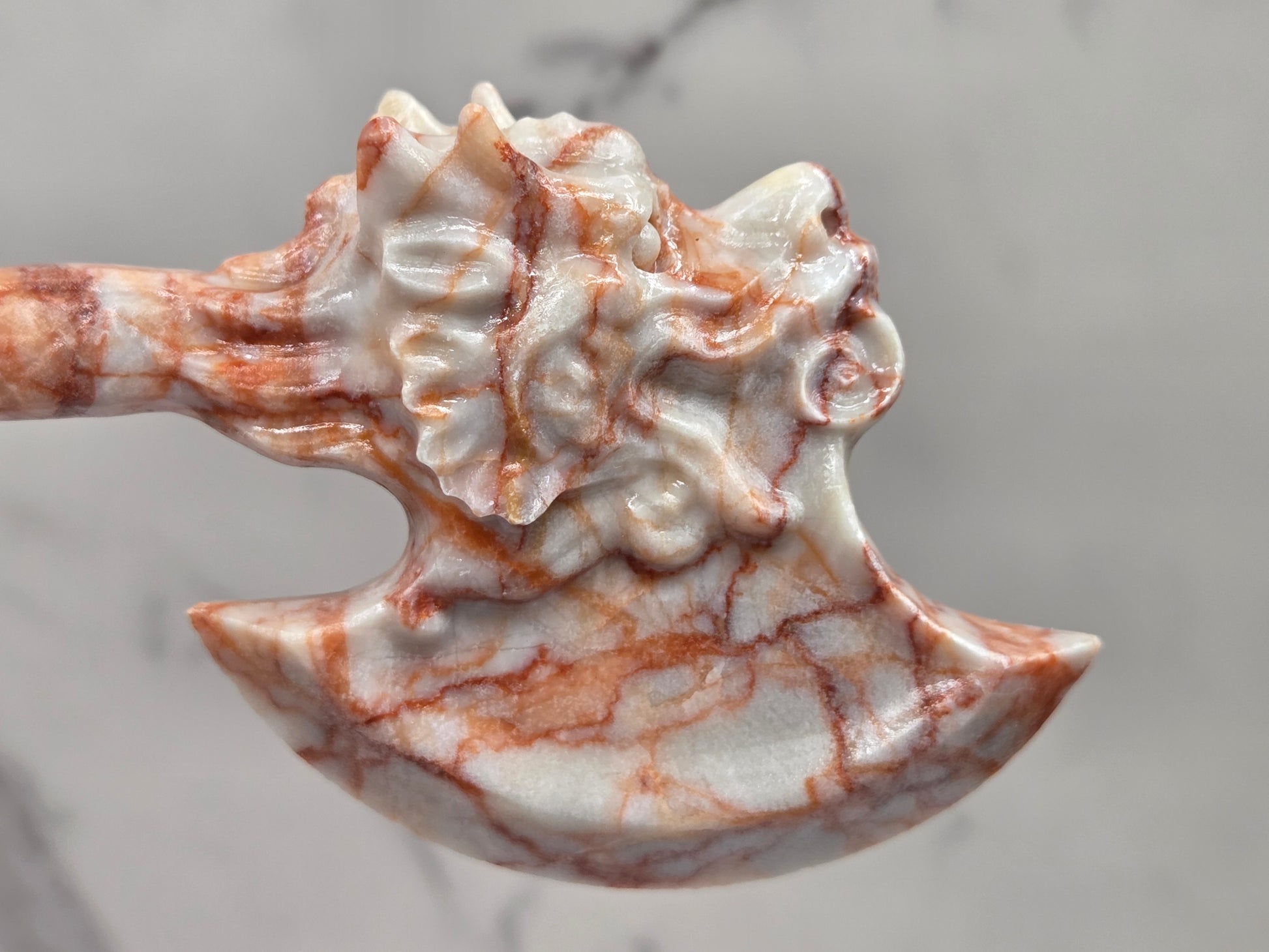 A close-up of a carved decorative axe or hatchet made from polished stone with a white base and reddish-brown marbling, set against a light gray marble-like background.