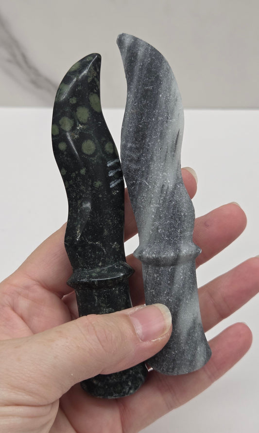 A hand holding two small handcrafted knives made from dark stone, with one knife open and the other closed, against a white and gray marble-patterned background.