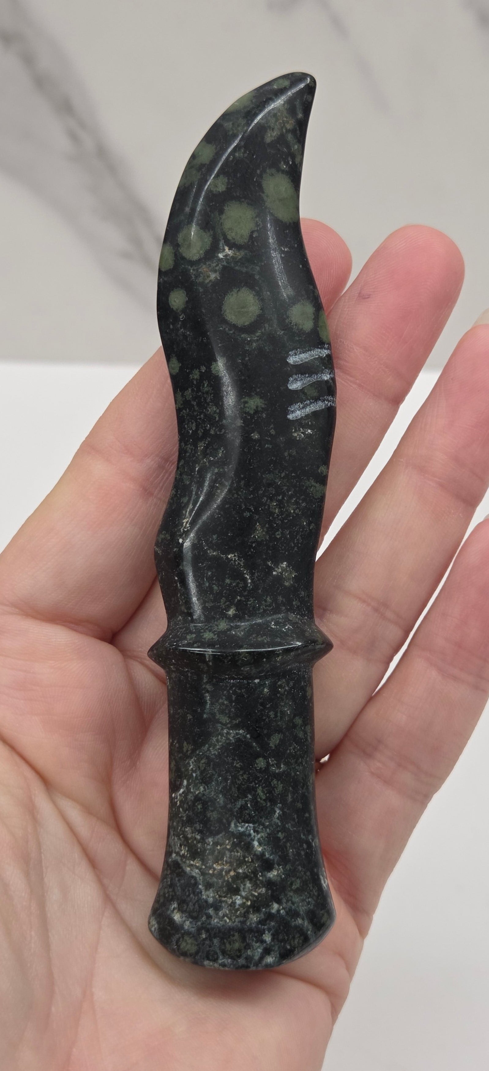 A hand holding a small polished black stone knife with green and white marbled inclusions, set against a light gray marble tile background.