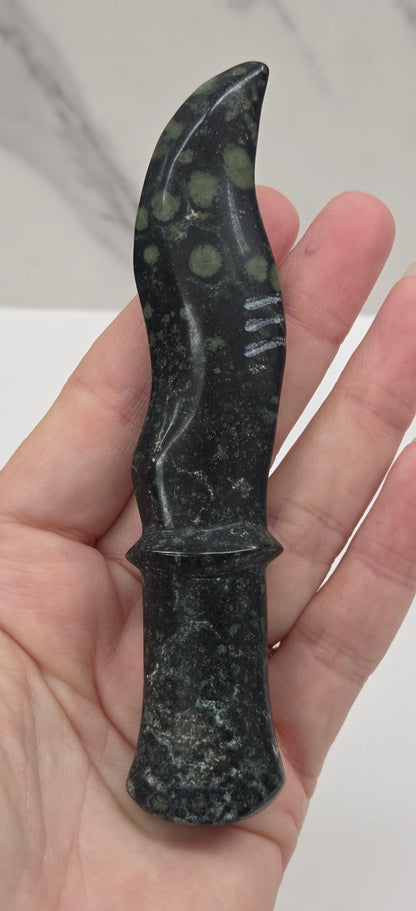 A hand holding a small polished black stone knife with green and white marbled inclusions, set against a light gray marble tile background.