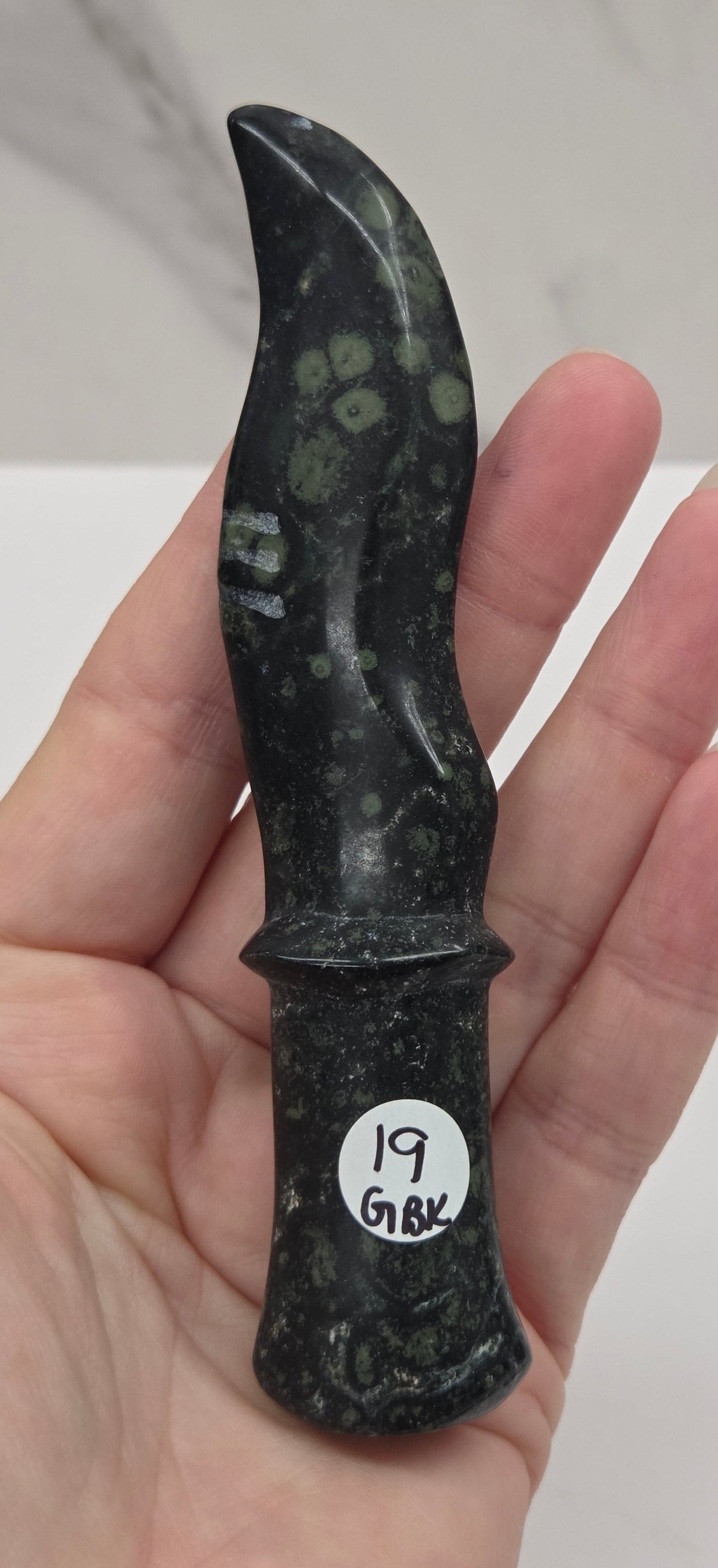 A hand holding a small black polished stone knife with natural inclusions, featuring a white circular sticker with handwritten text on the handle.