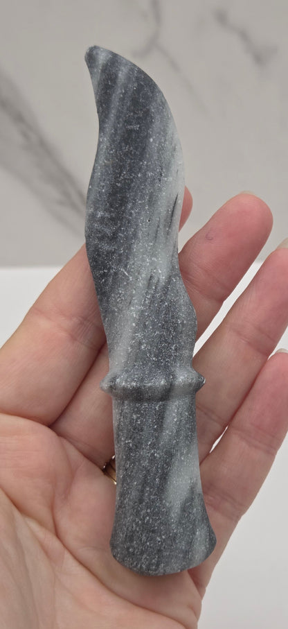 A hand holding a small gray stone knife with a textured, speckled finish against a light gray marble-patterned background.
