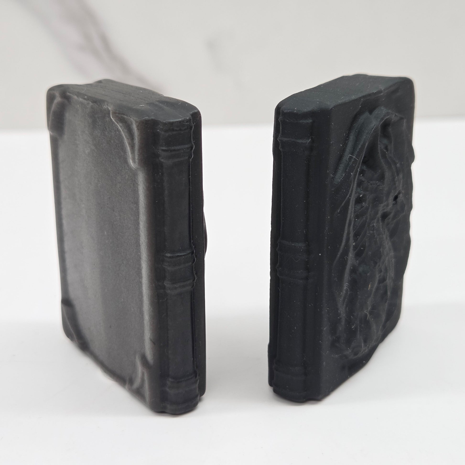 Two black, book-shaped carvings with textured, embossed details, standing upright on a white surface against a light gray background.