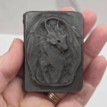 A hand holding a rectangular dark gray carved stone or resin plaque featuring a raised dragon design, set against a light marble background.
