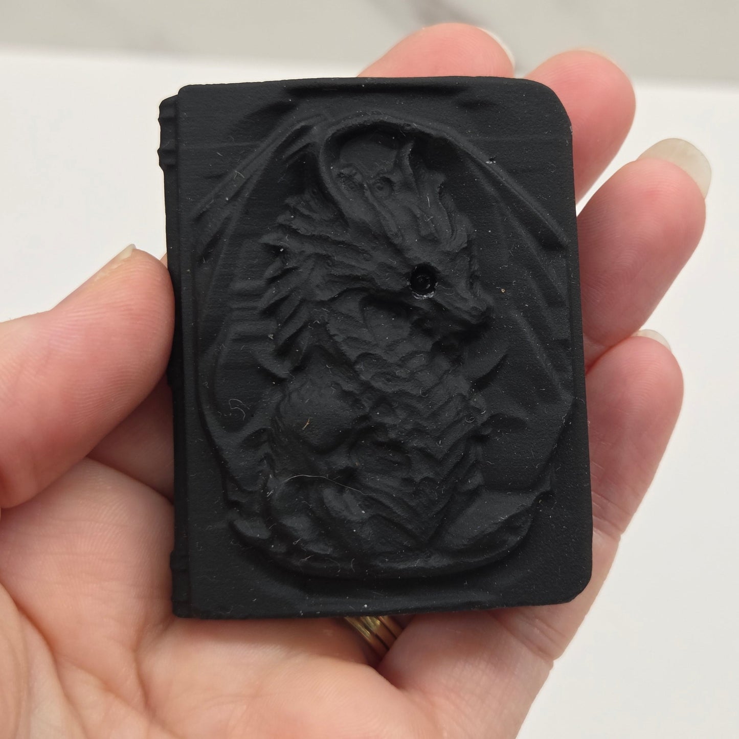A hand holding a black rectangular obsidian carving with a raised, textured design on its face, photographed against a light neutral background.