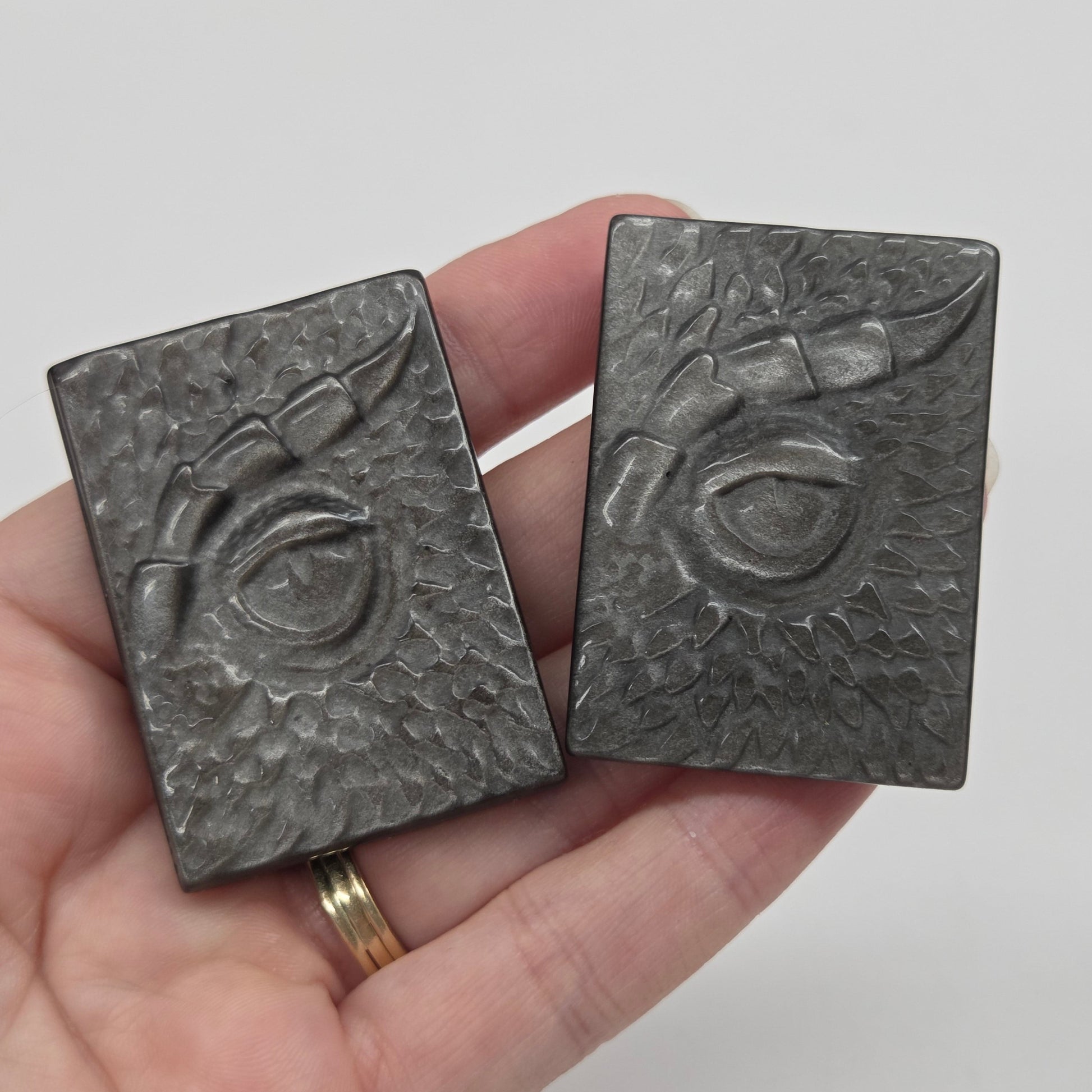 A hand holding two dark gray rectangular metal charms with embossed floral and circular designs, against a light gray background.
