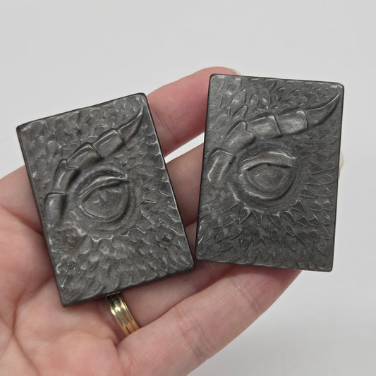 A hand holding two dark gray rectangular metal charms with embossed floral and circular designs, against a light gray background.
