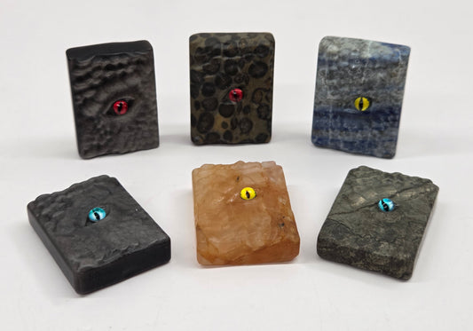 Six small rectangular stone or resin pendants with eye motifs are arranged on a white background. Four are dark charcoal/black with red, blue, or yellow eyes, and one is tan with a yellow eye.